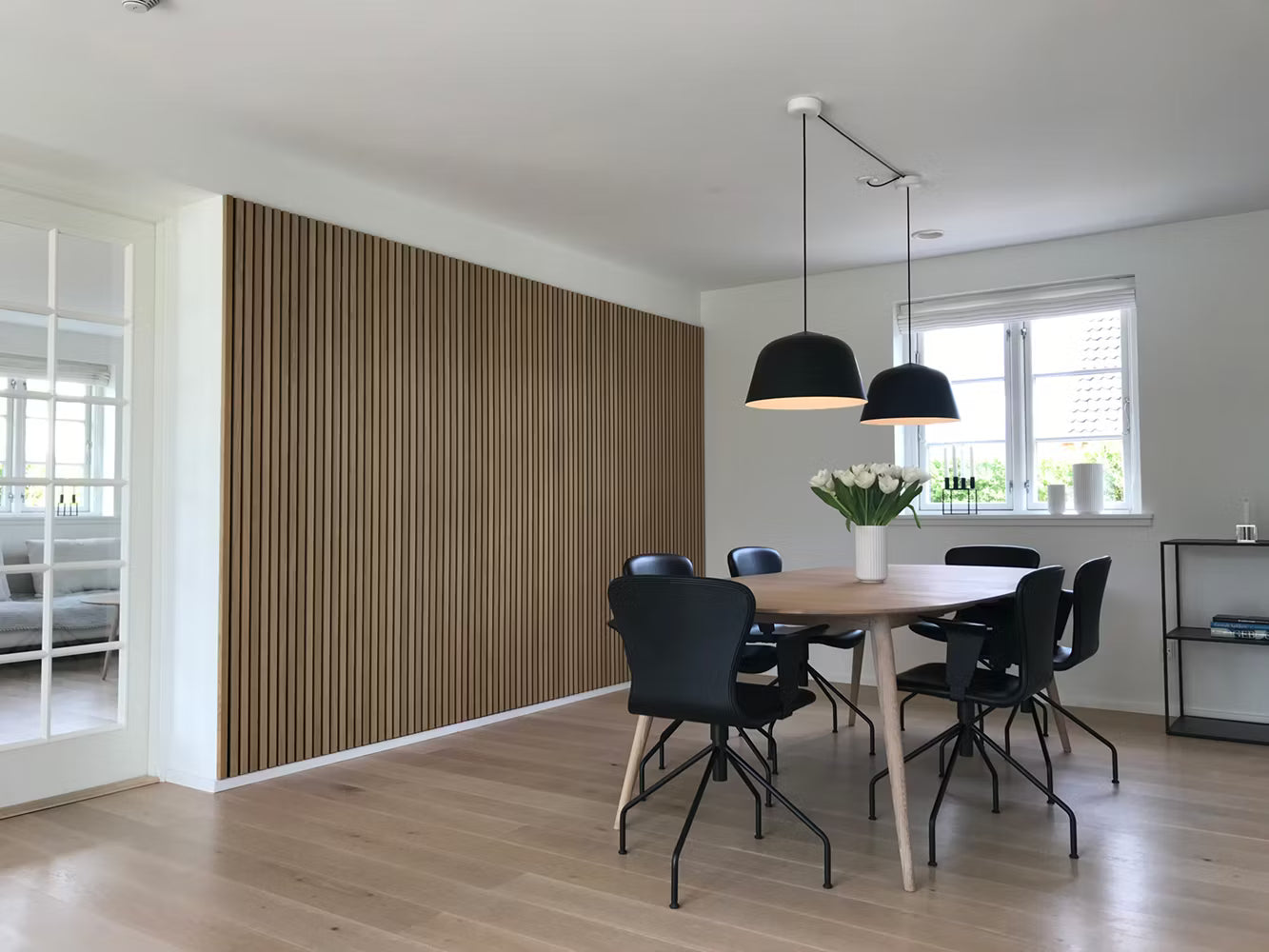 Nowoczesna jadalnia z drewnianą podłogą, akustycznym akcentem z listew Akupanel, czarnymi krzesłami, drewnianym stołem, lampami wiszącymi i białym kwiatowym elementem centralnym.
